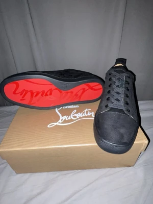 Svarta Christian Louboutin skor - Säljer ett par svarta sneakers från Christian Louboutin i mocka med klassisk röd sula och diskret logga på sidan. Skorna har rund tå, snörning och svart yttermaterial. Insidan är beige med guldfärgad logotyp. Perfekta för dig som vill ha något exklusivt och stilrent.