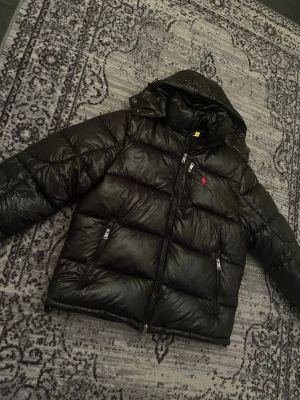 Svart pufferjacka från Polo Ralph Lauren - Säljer en svart pufferjacka från Polo Ralph Lauren med huva och flera dragkedjeförsedda fickor. Jackan har det klassiska röda Polo-logot på bröstet och är tillverkad i glansig polyester. Perfekt för kalla dagar och riktigt snygg streetstil.