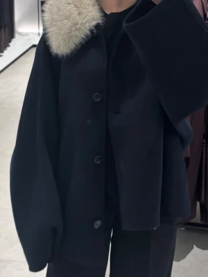 mörkblå ulljacka med fuskpäls från Zara - Säljer en mörkblå, kappa i ull med breda ärmar och stora knappar framtill. Kragen är i mjuk, ljus fuskpäls som ger en lyxig vibe. Perfekt för dig som vill ha en statementjacka till höst och vinter. Enkel men ändå riktigt snygg och trendig. Ser stor ut på mig då jag är 152cm!
