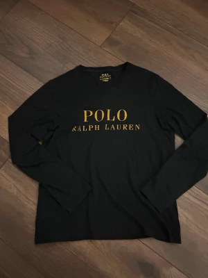 Svart långarmad från Ralph Lauren  - Svart långärmad topp från Polo Ralph Lauren med klassisk rund hals och stort gult logotryck framtill. Tillverkad i mjuk bomull som känns skön mot huden. Perfekt för dig som gillar stilrena och tidlösa plagg med en sportig touch.