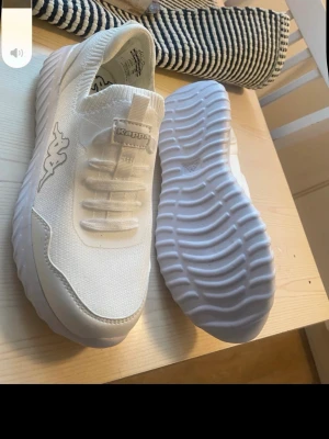 Vita sneakers från Kappa - Snygga vita sneakers från Kappa med klassisk logga på sidan. Skorna har en chunky, vågformad sula och ovandel i mesh och syntet. Elastisk snörning för enkel påtagning och sportig look. Perfekta för dig som gillar stilrena och bekväma skor.