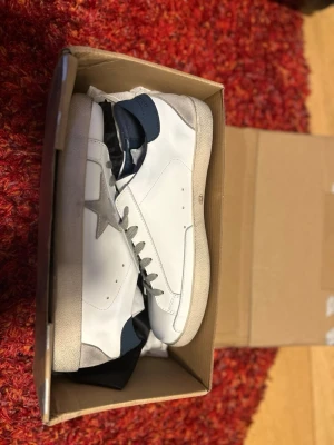 GGDB vita sneakers med grå stjärna - Säljer ett par GGDB sneakers i vitt skinn med grå mockadetaljer och en ikonisk stjärna på sidan. Skorna har grå snörning, blå hälflik och vit sula. Kommer med originalpåse och box. Perfekta för dig som gillar stilrena men ändå unika sneakers.