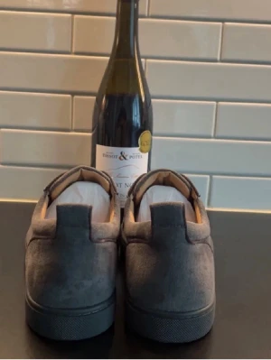 Gråa Louboutin sneakers med röd sula - Säljer ett par gråa sneakers från Louboutin med ikonisk röd sula och snygg snörning. Skorna är i mocka och har en stilren, rund tå och platt sula. Perfekta för dig som vill sticka ut med en lyxig touch och klassisk design. Pris kan sänkas vid snabb affär