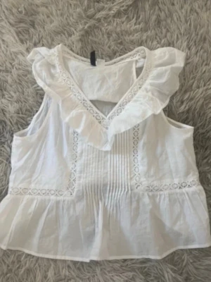 Vit blus med volang och spetsdetaljer - Söt vit blus med volang runt ringningen och spetsdetaljer längs framstycket. Blusen är ärmlös och har en luftig passform med fina veck framtill. Perfekt för varma dagar och ger en romantisk vibe till din outfit.