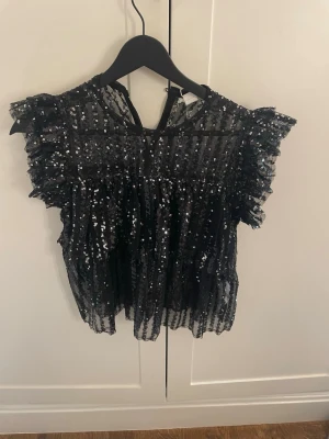 Svart paljettopp med eleganta volangärmar - Elegant svart paljettopp i lätt transparent mesh med vertikala paljettdetaljer som ger ett exklusivt och festligt uttryck. Toppen har korta volangärmar som skapar en modern och feminin silhuett. Bekväm passform och lätt croppad modell som passar perfekt till kjol, byxor eller jeans. Idealisk för fest, kalas och andra speciella tillfällen. Ny med etikett. 