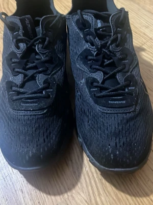 Svarta Nike React sneakers - Säljer ett par helsvarta Nike React sneakers med mesh-överdel och tydlig Nike-logga på sidan. Skorna har snörning, chunky sula och sportig look. Perfekta för dig som gillar stilrena och bekväma sneakers med modern design.