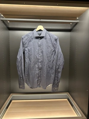 Mönstrad blå skjorta från Riley - Snygg blå skjorta från Riley med vitt geometriskt mönster och klassisk krage. Slim fit-modell med långa ärmar och vita knappar framtill. Perfekt för dig som gillar stilrena detaljer och modern passform.