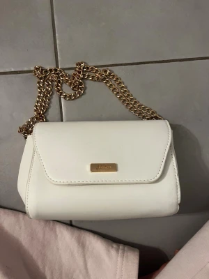 Vit axelväska med guldkedja  - Snygg vit axelväska från Sinsay med guldfärgad kedja och stilren design. Väskan har ett elegant metallmärke framtill och är gjord i slätt syntetmaterial. Kedjan går att justera. Inga defekter så den endast är använd 2 gånger. 