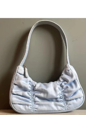 Handväska - !!!INTRESSEKOLL!!! Supersöt ljusblå axelväska från Monki i glansigt satinmaterial. Väskan har rynkade detaljer på framsidan och en praktisk dragkedja upptill. Insidan är fodrad och har ett extra fack för småsaker. Väskan ser mer babyblå ut än på bilderna. Den är lite nopprig🩵🩵