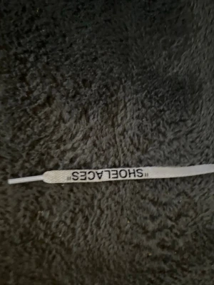 Vita skosnören med 'SHOELACES'-text - Säljer ett par vita skosnören med svart 'SHOELACES'-text, perfekta för att ge dina sneakers en unik Off-White-inspirerad vibe. Platta och i syntetmaterial, passar till de flesta sneakers och streetwear-stilar.