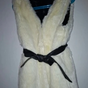 Vit pälsväst med svart bälte - Snygg vit pälsväst med mjuk och fluffig känsla, perfekt för att lyfta din outfit. Västen har ett svart bälte i midjan som knyts för att ge en cool siluett. Passar dig som gillar att sticka ut med stilrena detaljer.