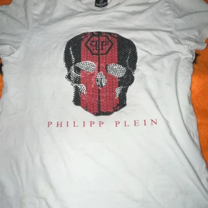 ÄKTA! Vit qp tröja 164 passar även 150 - Snygg vit t-shirt från Philipp Plein med ett stort döskalletryck i svart och rött framtill. T-shirten har korta ärmar och rund hals. Märkesnamnet är tryckt i rött under motivet. Perfekt för dig som gillar edgy streetstyle.
