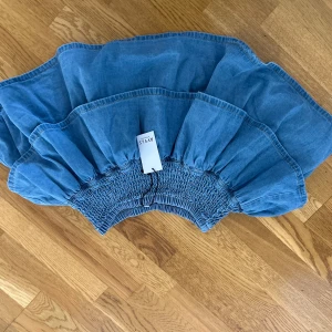 Blå volangkjol i denim från RYVLS - Supersöt blå kort volangkjol i denim från RYVLS. Kjolen har smockad midja och flera lager med volanger som ger en riktigt snygg och trendig look. Perfekt att matcha med en enkel topp eller hoodie för en chill stil.