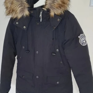 Fin parkasjacka med fuskpäls från Svea - Fin parkasjacka från Svea med fuskpäls på huvan och emblem på ärmen. Jackan har dragkedja och knappar. Perfekt för kalla dagar och har en klassisk, tidlös look.