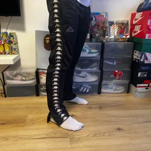 Svarta Kappa trackpants med logoband - Svarta trackpants från Kappa med ikoniska vita logoband längs båda sidorna. Byxorna har elastisk midja och tryckknappar längs benen för en retro vibe. Materialet är polyester och passformen är loose. Perfekt för dig som gillar streetwear och sportig stil.