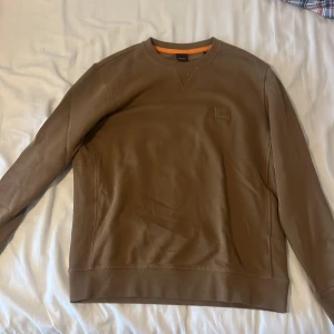 Beige sweatshirt från BOSS - Säljer en stilren beige sweatshirt från BOSS med rund halsringning och diskret logopatch på bröstet. Tröjan har långa ärmar och ribbade muddar vid ärmslut och nederkant. Perfekt för en clean och avslappnad look.