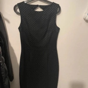 Svart ärmlös kortklänning med mönster - Snygg svart ärmlös klänning med diskret rutmönster och figurnära passform. Klänningen har en elegant detalj i nacken och är perfekt för dig som gillar stilrena plagg med det lilla extra. Passar dig som vill ha en klassisk look med modern twist.