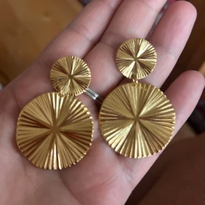 guldiga örhängen  - Säljer ett par runda statementörhängen i guld med snyggt räfflat solstrålemönster. Örhängena har två cirklar i olika storlekar som sitter ihop och ger en cool, iögonfallande look. Perfekta för dig som vill sticka ut och addera lite extra bling till din outfit. Dem liknar Caroline Svedbom örhängen modell Melia