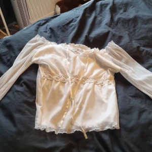 Vit blus med spetsdetaljer - Romantisk vit blus med långa transparenta ärmar och spetsdetaljer vid midjan och ärmsluten. Blusen har knytband framtill och volangkant längs halsringningen och nederkanten. Perfekt för dig som gillar en söt och bohemisk stil.