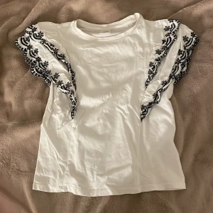 Vit blus med broderade volangärmar Zara - Supersöt vit blus från Zara med korta volangärmar som har svart broderi i blommigt mönster. Toppen är i mjuk bomull och har en klassisk rund halsringning. 