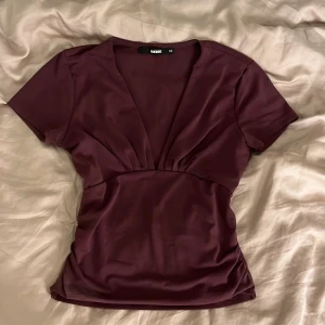 Vinröd v-ringad topp från BikBok - Säljer en vinröd topp från BikBok i storlek XS. Toppen har djup v-ringning, korta ärmar och smickrande rynkdetalj under bysten. Materialet är mjukt och stretchigt, perfekt för en snygg och bekväm look.