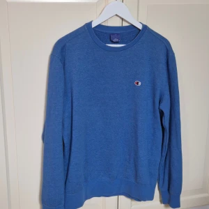 Blå vintage crewneck från Champion L - En klassisk blå vintage crewneck från Champion med broderad logga på bröstet. Tröjan har rund halsringning, långa ärmar och ribbade muddar. Perfekt för dig som gillar en enkel och stilren look. 