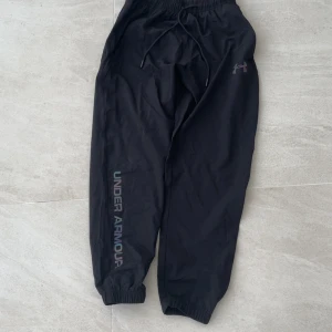 Svarta träningsbyxor från Under Armour - Svarta träningsbyxor från Under Armour med regnbågsfärgat logotryck på benet och logga på höften. Byxorna har resår och snörning i midjan samt resår vid bensluten. Perfekta för dig som gillar sportig och bekväm stil.