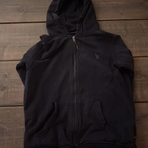 Svart hoodie från Polo Ralph Lauren - Svart hoodie från Polo Ralph Lauren med dragkedja framtill och klassisk huva. Tröjan har två fickor på magen och en diskret broderad logga på bröstet. Perfekt för lager-på-lager och chill dagar. Tillverkad i mjuk bomull för extra komfort.