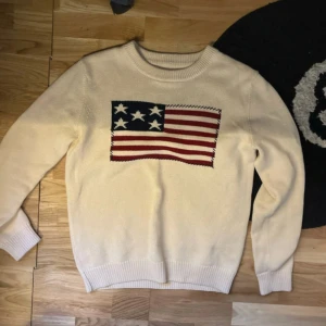 Beige stickad tröja Selected Homme - Snygg beige stickad tröja från Selected Homme med amerikansk flagga i blått, vitt och rött på bröstet. Rund hals och ribbade muddar vid ärmslut och nederkant. Perfekt för dig som vill ha en unik och cool look.
