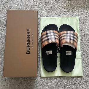 Burberry tofflor/sandaler rutig beige/svart - Säljer ett par snygga Burberry sandaler med klassiskt rutigt mönster i beige, svart, vitt och rött. De har en platt svart sula och bred rem över foten i syntetmaterial. Perfekt för dig som vill ha en lyxig och stilren look.