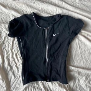 Svart kortärmad topp från Nike - Säljer en svart kortärmad topp från Nike med dragkedja framtill och vit piping längs kanterna. Perfekt för en sportig look eller avslappnad stil. Nike-loggan är diskret placerad på bröstet.