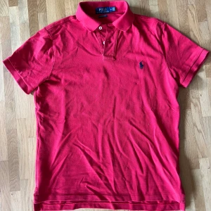 Röd pikétröja från Polo Ralph Lauren - Säljer en röd pikétröja från Polo Ralph Lauren i modellen Custom Slim Fit. Tröjan har klassisk krage, korta ärmar och knappar framtill. Ikoniska loggan broderad på bröstet. Perfekt för dig som gillar stilren och sportig look.