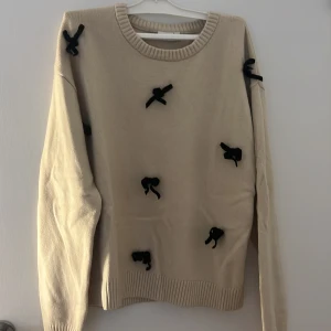 Beige stickad tröja med svarta rosetter - Superfin beige stickad tröja från name it med svarta rosetter som detaljer. Tröjan har rund halsringning, långa ärmar och ribbade muddar. Perfekt för dig som vill ha något gulligt och unikt i garderoben!