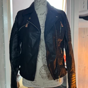 Svart skinnjacka från Voyelles - Snygg svart skinnjacka från Voyelles med coola detaljer som dragkedjor och quiltade partier på ärmarna. Jackan har klassisk bikerstil med snedställd dragkedja och flera fickor framtill. Perfekt för dig som vill ha en edgy look. Liten i storlek