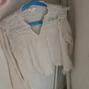 Vit romantisk blus med volang - Supersöt vit blus med volangdetaljer och v-ringning. Blusen har långa ärmar och fina spetsinslag upptill som ger en romantisk känsla. Perfekt att styla med jeans eller kjol för en trendig look.