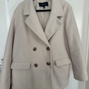 Beige kappa  - Säljer en beige fin kappa från Vero Moda med dubbelknäppning och stora, mönstrade knappar. Liten fläck (se bild 3) men går att få bort, annars i nyskick