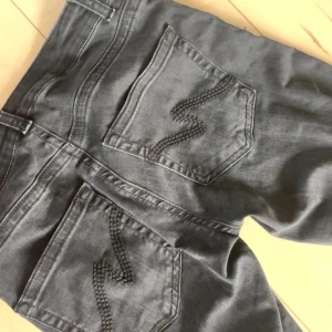 Nelly jeans  - Säljer ett par svarta jeans med snygga slitningar på båda knäna och coola broderade detaljer på bakfickorna. Jeansen har rak passform och klassisk femficksdesign. Perfekta för dig som gillar en avslappnad och trendig stil.