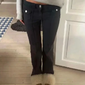 Svarta bootcut byxor med knappar - Snygga svarta byxor med bootcut-modell och dekorativa knappar framtill. Byxorna har låg midja och markerade pressveck längs benen för en clean look. Perfekta att styla med crop top eller hoodie.