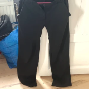 Svarta kostymbyxor med rak passform - Snygga svarta kostymbyxor med klassisk rak passform och knappstängning fram. Perfekta för en clean och stilren look. Materialet känns mjukt och har en lätt stretch, vilket gör dem bekväma att bära hela dagen.