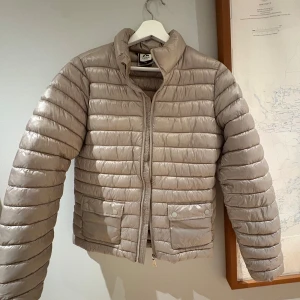 Beige pufferjacka från McKinley - Snygg beige pufferjacka från McKinley med horisontella sömmar, hög krage och två fickor framtill med tryckknappar. Jackan har dragkedja och är lätt vadderad, perfekt för kyliga dagar. Enkel och stilren design som passar till det mesta.