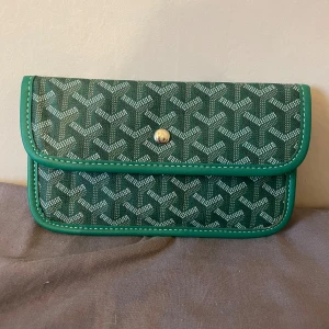 Grön kuvertväska med mönster - Snygg grön kuvertväska med grafiska mönster. Väskan har silverfärgad knappstängning, gröna kanter och insida i ljust tyg. Perfekt för att bära det viktigaste och ge din outfit en lyxig touch. Det är ett märke inne i väskan men annars i jättebra skick