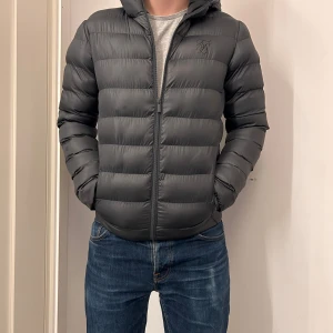 Mörkgrå pufferjacka från SikSilk XS - Snygg mörkgrå pufferjacka från SikSilk i storlek XS. Jackan har huva, raka kanaler och dragkedja framtill. Perfekt för kalla dagar och har en stilren look med diskret logga på bröstet. Materialet är syntet och jackan är långärmad.