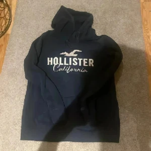 Svart hoodie från Hollister California - Snygg mörkblå hoodie från Hollister med stort vitt tryck på bröstet och klassisk logga. Tröjan har huva med snörning och en skön, mjuk insida. Perfekt för dig som gillar en avslappnad och stilren look. Strl L men liten så passar fint på s/m lite oversized❤️