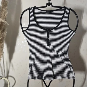 Randig linnetopp från Lindex - Snygg svartvit randig linnetopp från Lindex i storlek S. Toppen har rund halsringning och knappar framtill som detalj. Tillverkad i mjuk bomull med smal passform och ärmlös design. Perfekt för dig som gillar enkel och stilren look.