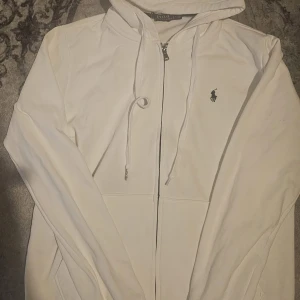 Vit hoodie från Polo Ralph Lauren - Vit hoodie från Polo Ralph Lauren med dragkedja och huva. Klassisk broderad logga i svart på bröstet och snörning vid huvan. Mjuk bomullskvalitet och långärmad modell, perfekt för chill dagar. Snygg och enkel design som funkar till allt.