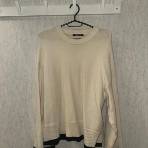 Beige stickad tröja från Gina Tricot - Snygg beige stickad tröja från Gina Tricot med svarta detaljer vid ärmslut och nederkant. Tröjan har en loose passform, rund hals och långa ärmar med breda ribbade muddar. Inget att anmärka. Storlek M