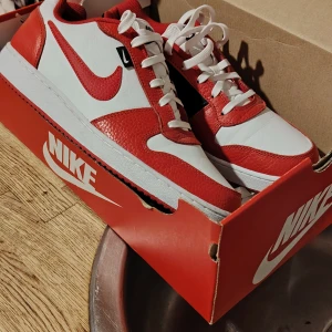 Nike Air Force 1 röd/vit sneakers i kartong - Nike Air Force 1 sneakers i en snygg röd och vit färgkombination. Skorna har klassisk låg siluett, rund tå och platt sula. Tillverkade i skinn med vita snören och ikonisk röd Swoosh på sidan. Perfekta för dig som vill sticka ut med stil.