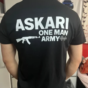 Svart ASKARI t-shirt med tryck - Svart t-shirt från ASKARI med vit text och motiv på både bröstet och ryggen. Stort tryck på ryggen med texten 'ASKARI ONE MAN ARMY' och vapenmotiv. Klassisk passform och rund hals. Perfekt för dig som gillar streetwear med attityd.