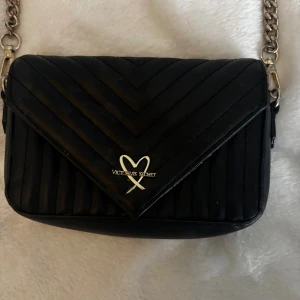 Svart victoria secret väska - Snygg svart axelväska från Victoria's Secret med quiltad design och guldfärgad kedja. Väskan har ett hjärtformat metallmärke framtill och ett stilrent, envelope-format lock. Perfekt storlek för det viktigaste och ger en cool vibe till din outfit.