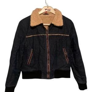 Mörkblå jeansjacka med teddykrage - Snygg mörkblå jeansjacka med mysig beige teddykrage och detaljer i samma material längs bröstet. Jackan har dragkedja framtill, ribbade muddar och två fickor med dragkedja. Perfekt för dig som vill ha en cool och bekväm look.